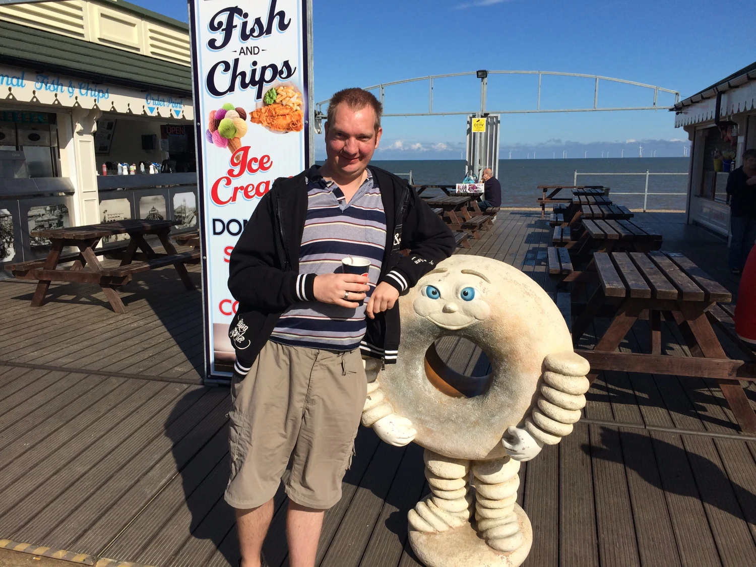 A man posing beside a sheep figure on a seafront promenade